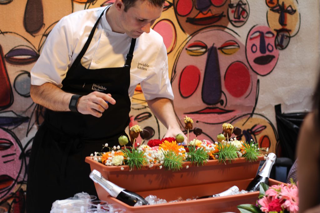 chef tom infront of a wall with faces painted on with colorful food and flowers